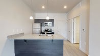 This single-floor residence showcases a contemporary design with a clean, neutral color palette throughout. The open-concept kitchen serves as a central highlight, anchored by a large island with granite countertops and an integrated sink. Dark-toned cabinetry provides strong visual contrast against the white walls, while a full suite of stainless steel appliances — including a refrigerator, electric range, and over-the-range microwave — delivers a polished, functional finish. Pendant lighting hangs above the island, adding warmth and definition to the space. Light wood-style flooring extends through the kitchen and common areas, contributing to a bright and cohesive aesthetic.

The bedroom features plush carpeting and neutral wall tones, with a window fitted with horizontal blinds that allows natural light and outdoor views of surrounding greenery. The bathroom is well-appointed with a granite-topped vanity, a full tub-shower combination, light wood-style flooring, and a conveniently integrated stacked washer and dryer unit. Overall, the space presents a modern, move-in-ready environment with consistent finishes and thoughtful functionality throughout.