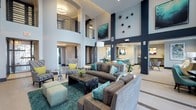 This contemporary communal lounge is defined by its dramatic double-height ceilings, which lend an expansive, airy quality to the space. Tall navy blue structural columns rise prominently throughout, providing strong architectural contrast against the crisp white walls and light-toned flooring. An upper-level mezzanine with sleek metal railings overlooks the main lounge area below, adding architectural depth and visual interest.

The seating area is thoughtfully arranged around a dark, textured coffee table set atop a rich teal area rug. Plush grey sofas are styled with teal and navy accent pillows, while patterned armchairs feature bright yellow cushions, creating a lively, coordinated color palette throughout. A sculptural arc floor lamp provides ambient lighting, complementing the recessed ceiling lights above.

Wall décor includes a bold abstract artwork framed prominently on one column, alongside a striking cluster of decorative black sunburst accents on an adjacent wall. In the background, a dining or bar area with teal seating is visible, suggesting additional functional zones within the space. The open layout and refined design details combine to create a polished and inviting communal environment.