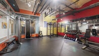 This energetic fitness facility is divided into two distinct workout zones, each designed to support a comprehensive training experience. The main strength training area features an open, industrial aesthetic with exposed ductwork and piping along the ceiling, track lighting, and bold accent walls in red, orange, and yellow. This space is outfitted with a cable machine, multiple power racks, and a full dumbbell rack, all set on durable rubber flooring. Large mirrors line the walls to maximize the sense of space and support proper form during workouts.

The cardio zone offers a more intimate setting, distinguished by a striking striped mural in red, yellow, and orange hues, complemented by a bold star graphic as a focal point. This area houses multiple treadmills, an elliptical, a stair climber, and a stationary bike, each equipped with individual monitors. A wall-mounted television provides entertainment during sessions. Stability balls and other functional training accessories are also available. The dark ceiling and recessed track lighting in this zone create a focused, energizing atmosphere. Together, both areas provide a well-rounded and motivating environment suited for a variety of fitness routines.