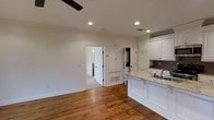 This single-story space showcases a seamlessly connected kitchen and living area, unified by warm-toned hardwood floors that run throughout. The kitchen is outfitted with white raised-panel cabinetry, offering generous storage, and is complemented by light granite countertops with subtle veining. A peninsula with an undermount sink provides additional workspace and serves as a natural divider between the kitchen and the open living area. A tile backsplash adds a tasteful accent behind the counters. Recessed lighting is installed throughout the ceiling, providing consistent, even illumination across the space, while a ceiling fan in the living area contributes to both comfort and airflow. Crown molding along the ceiling adds a classic architectural detail. A doorway leads to an adjacent room, suggesting a practical and connected floor plan. Built-in cabinetry near the transition area offers additional storage solutions. The neutral, light-toned walls enhance the brightness of the space, creating a clean and welcoming atmosphere overall.