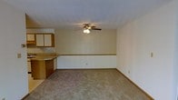 This single-floor residence offers a straightforward and functional layout designed for comfortable everyday living. The open-plan living area is finished with neutral wall-to-wall carpeting that lends a warm, understated feel to the space. A ceiling fan with an integrated light fixture serves as the central overhead lighting source, providing both illumination and air circulation throughout the room. A decorative chair rail runs along the perimeter walls, adding a subtle architectural detail that breaks up the neutral wall surfaces. The kitchen is positioned adjacent to the living area and features warm-toned oak cabinetry, offering ample storage space. A countertop peninsula or breakfast bar creates a natural transition between the kitchen and living space, making it well-suited for casual dining or food preparation. The cabinetry extends to the upper wall, maximizing vertical storage. The overall ambiance is clean, neutral, and move-in ready, with a cohesive color palette of beige and warm wood tones throughout. The single-floor design ensures easy accessibility and a seamless flow between living and kitchen areas, making the space practical and welcoming.
