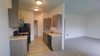 This single-floor space showcases a well-designed open-concept layout where the kitchen flows naturally into the adjoining living area. The kitchen is outfitted with contemporary medium-toned wood cabinetry, both upper and lower, providing ample storage throughout. Light-colored countertops offer a clean contrast against the cabinetry, while a stainless steel sink is positioned along the counter with a practical layout. Black appliances, including a refrigerator, stove, and dishwasher, add a bold accent to the kitchen's neutral palette. Light hardwood-style flooring runs through the kitchen zone, transitioning into soft carpeting in the living area, creating a clear and comfortable delineation between spaces. Flush-mount ceiling fixtures provide consistent overhead lighting throughout. A hallway visible in the background leads to additional areas of the unit, including what appears to be a closet with shelving. The overall color scheme is kept neutral with light walls and warm wood tones, contributing to a bright and clean aesthetic throughout the space.
