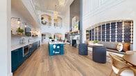 Located at 5315 Waters Edge Village Drive in Charlotte, NC, this impressively scaled community clubhouse at Novel Waters Edge delivers a thoughtfully designed, open-concept gathering space that spans nearly 2,000 square feet on a single floor. The interior is anchored by dramatic double-height ceilings that rise above the main social hall, enhanced by elegant chandelier lighting and an upper mezzanine railing that adds architectural depth. Wide-plank hardwood flooring flows throughout, unifying the space with warmth and continuity. A bold, floor-to-ceiling dark shiplap fireplace wall serves as a striking focal point, flanked by built-in shelving and a wood beam mantle. Adjacent to the fireplace is a stylishly appointed lounge area with a distinctive geometric-patterned accent wall. The open kitchen area features deep teal cabinetry, a white quartz island with bar seating, and premium finishes. Large black-framed floor-to-ceiling windows and glass doors flood the interior with natural light while offering views of the surrounding outdoor spaces. A dedicated game and recreation area near the entrance provides additional social functionality. A separate lounge room with a bold patterned ceiling, sheer drapery, and ample seating creates a refined retreat. Throughout, the design balances contemporary sophistication with welcoming, residential warmth.