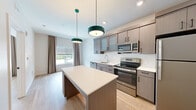 Located at 8895 Perimeter Park Blvd in Jacksonville, FL, this one-bedroom, one-bathroom unit offers a thoughtfully designed layout that maximizes every square foot. The open-concept kitchen serves as the centerpiece of the living area, featuring gray wood-tone cabinetry with both frosted glass and solid upper cabinet doors, sleek white quartz countertops, and a full suite of stainless steel appliances including a refrigerator, electric range, over-the-range microwave, and dishwasher. A substantial kitchen island with built-in shelving provides additional storage and workspace, while a pair of teal pendant lights adds a modern, distinctive touch. Light wood-tone flooring flows consistently throughout the unit, contributing to a clean and cohesive aesthetic. The bedroom offers a comfortable and well-lit retreat, with a large window dressed in neutral drape panels that invite natural light and outdoor views. The bathroom is finished to a high standard, showcasing a gray vanity with white countertop, drawer storage, open shelving, an LED-lit mirror, and adjacent built-in open shelving for added organization. Crisp white walls and consistent finishes create a bright, contemporary ambiance throughout this well-appointed Jacksonville unit.