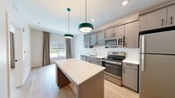 Located at 8895 Perimeter Park Blvd in Jacksonville, FL, this one-bedroom, one-bathroom unit offers a thoughtfully designed layout that maximizes every square foot. The open-concept kitchen serves as the centerpiece of the living area, featuring gray wood-tone cabinetry with both frosted glass and solid upper cabinet doors, sleek white quartz countertops, and a full suite of stainless steel appliances including a refrigerator, electric range, over-the-range microwave, and dishwasher. A substantial kitchen island with built-in shelving provides additional storage and workspace, while a pair of teal pendant lights adds a modern, distinctive touch. Light wood-tone flooring flows consistently throughout the unit, contributing to a clean and cohesive aesthetic. The bedroom offers a comfortable and well-lit retreat, with a large window dressed in neutral drape panels that invite natural light and outdoor views. The bathroom is finished to a high standard, showcasing a gray vanity with white countertop, drawer storage, open shelving, an LED-lit mirror, and adjacent built-in open shelving for added organization. Crisp white walls and consistent finishes create a bright, contemporary ambiance throughout this well-appointed Jacksonville unit.