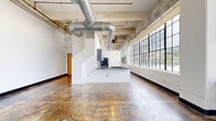 Located at 820-828 Ralph McGill Blvd NE in Atlanta, GA, this single-floor industrial loft space delivers an open, airy atmosphere defined by its raw architectural character. The main area is expansive and column-accented, showcasing polished concrete floors with a rich, variegated patina that adds warmth and texture to the industrial aesthetic. Soaring ceilings with exposed concrete soffits and prominent galvanized HVAC ductwork run the length of the space, reinforcing its converted warehouse character. Large steel-framed, multi-pane windows line the walls, flooding the interior with abundant natural light and offering views of the surrounding Atlanta skyline and greenery. White-painted cinder block walls provide a clean contrast to the raw structural elements overhead. A built-in kitchen counter with a sink serves as a functional anchor point within the open floor plan. The hallway connects to a bathroom featuring dark slate-tiled shower walls and earthy floor tiles, lending a bold, contrasting finish to the utilitarian-inspired design. A ceiling fan and wall-mounted HVAC unit provide climate control throughout. The overall ambiance is one of creative versatility — an industrial canvas well-suited for a variety of uses in the heart of Atlanta.