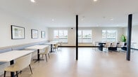 Located at 3980 Shelbourne St in Saanich, BC, this single-floor communal space offers a well-appointed, open-plan living area designed for comfort and functionality. The interior is defined by light hardwood-style flooring that runs throughout, crisp white walls, and recessed ceiling lighting that ensures a bright, welcoming atmosphere.

The main area is generously proportioned and organized into distinct seating zones. A built-in banquette along one wall is paired with a series of white rectangular tables and modern chairs, creating a comfortable lounge-dining arrangement. A row of framed artwork in neutral tones lines the wall above the banquette, adding a refined aesthetic touch. A second grouping of seating features a round dark table surrounded by contemporary chairs, offering a more intimate setting.

Bold black structural columns punctuate the open floor plan, contributing an industrial design element that contrasts effectively with the clean, minimal decor. A separate alcove provides a quieter retreat, furnished with two swivel chairs with red accent cushions and a low round coffee table, ideal for private conversation.

The entrance features large glazed panels that introduce natural light, complemented by a tall potted fiddle-leaf fig that adds a lively natural element. Multiple windows along the perimeter frame views of the surrounding neighbourhood, keeping the space feeling connected to the outdoors.