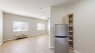 This single-story, one-bedroom, one-bathroom unit offers a practical and efficiently designed layout. The open living area is finished with light wood-style laminate flooring that runs throughout, creating a clean and cohesive look. Two windows fitted with horizontal blinds allow natural light to fill the space, complemented by a ceiling fan with a light fixture overhead. A wall-mounted HVAC unit provides climate control directly in the living area.

The kitchen is partially separated from the main living space by a partial wall, and is equipped with a stainless steel refrigerator, a range, and light wood cabinetry, along with an adjacent built-in shelving unit for additional storage.

The spacious primary bedroom features the same light laminate flooring, a ceiling fan, and a built-in closet with a standard rod and shelf system, offering ample storage.

The bathroom is designed with accessibility in mind, featuring a roll-in or walk-in shower with a handheld showerhead, stainless steel grab bars positioned along the walls, and a barrier-free entry. The neutral color palette of warm beige and white tones throughout the unit contributes to a bright, clean, and functional living environment.