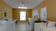 This single-floor laundry facility is designed with functionality and accessibility in mind. The room is finished with warm golden-yellow walls that create a welcoming atmosphere, complemented by light-toned tile flooring that is easy to maintain. Overhead, bright fluorescent lighting ensures the space is well-lit throughout.

The laundry room is equipped with multiple front-loading washers and top-loading dryers arranged along the walls, providing ample capacity for users. A folding counter is positioned along one wall, offering a convenient surface for sorting and folding laundry. A window along one wall allows natural light to supplement the overhead fixtures, adding a sense of openness to the space.

Instructional signage, including laundry usage guides and a LaundryView panel for monitoring machine availability, is posted on the walls, enhancing usability. Payment or control terminals are also visible near the machines. The room's straightforward layout maximizes floor space while keeping all equipment easily accessible, making it a practical and well-organized communal laundry facility.