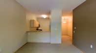 This single-floor residential unit offers a practical and comfortable layout designed for everyday living. The kitchen is outfitted with light wood cabinetry, a stone-topped breakfast bar, and essential appliances including a microwave, making it both functional and inviting. The peninsula-style counter creates a natural division between the kitchen and the adjoining living spaces while maintaining an open, connected feel throughout.

Neutral-toned walls and flush ceiling lighting keep the interior feeling bright and clean. The flooring transitions from the kitchen area into soft wall-to-wall carpeting in the living spaces, adding warmth underfoot. A partial wall column defines the transition between zones without fully closing off the floor plan, preserving the sense of openness.

Moving further into the unit, a pendant light fixture illuminates a secondary area — likely a dining nook or additional living space — adding a decorative touch to the otherwise understated interior. A door visible at the far end hints at access to additional rooms or building common areas. The overall ambiance is clean, neutral, and move-in ready, suited for those seeking a well-organized, low-maintenance living environment.