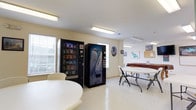 Located at 232 W. Gilbert St. in Hampton, Virginia, this single-floor facility offers a practical and welcoming environment designed for community use. The main gathering area is bright and open, illuminated by overhead fluorescent lighting and supplemented by natural light filtering through window blinds along the exterior walls. The space features light-colored walls and a clean, polished floor that contributes to the overall sense of openness.

The room is equipped with both a snack vending machine and a cold drinks vending machine, providing convenient refreshment options for occupants. Folding tables and chairs are arranged throughout the area, making it adaptable for a variety of uses including meetings, gatherings, or casual socializing. A cork bulletin board is mounted on the wall, serving as an informational hub for community notices and announcements. Decorative wall art, including nature-themed imagery, adds a modest visual interest to the space.

The layout is straightforward and functional, with ample room to move freely between seating arrangements. The overall ambiance is clean, utilitarian, and community-oriented, making this space well-suited for residents or groups seeking a shared, accessible area for everyday activities and organized events.