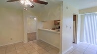 This single-story home showcases a practical and open layout that connects the main living areas seamlessly. The main living space features light-colored tile flooring and a vaulted ceiling with a notable exposed wood beam, adding architectural character to the room. A ceiling fan is mounted overhead, contributing to both style and comfort.

The kitchen is positioned behind a half-wall pass-through, offering an open connection to the living area while maintaining a defined cooking space. It is equipped with a white range, an overhead range hood, and wooden cabinetry in a warm medium-brown tone. A white refrigerator is also visible, and the countertops are light-colored, complementing the overall neutral palette of the home.

Adjacent to the main living area, a doorway leads into a carpeted room, suggesting a bedroom or secondary living space, also equipped with its own ceiling fan. The walls throughout are finished with a light green-tinted paint, and a textured wall surface adds subtle depth to the interior. The overall ambiance is bright, airy, and functional, offering a straightforward and well-connected floor plan.