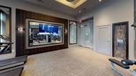 This fitness center offers a thoughtfully designed workout environment that balances functionality with contemporary aesthetics. The space is defined by floor-to-ceiling glass windows along one wall, flooding the interior with natural light and providing open views of the surrounding streetscape. The ceiling features an elegant tray design accented by circular pendant light fixtures and recessed lighting, contributing to a polished, upscale ambiance throughout.

The main workout area is equipped with a diverse array of cardio machines, including treadmills, ellipticals, and stationary bikes, all positioned to take advantage of the abundant natural light from the expansive glazed facade. A dedicated strength training zone includes a multi-station cable machine, a full power rack, adjustable benches, and a dumbbell rack, catering to a wide range of training needs.

A prominent large-format display screen is framed in rich wood paneling on one wall, serving both an entertainment and motivational function. Translucent glass partition panels define areas within the space while maintaining visual openness. The neutral-toned carpeted flooring complements the overall muted, sophisticated color palette, creating a focused and professional fitness environment.
