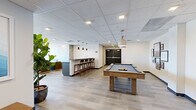 This generously sized lounge is thoughtfully designed to serve as a versatile social hub, offering a variety of activity and relaxation zones across its single open floor plan. Light wood-toned tile flooring runs throughout, lending warmth and visual continuity to the space.

A central wet bar with dark cabinetry, a white countertop, and pendant lighting serves as an anchor point, complemented by an adjacent high-top bar counter with barstool seating. A full-size pool table with a wood-frame base occupies a prominent position in the main activity area, while a shuffleboard table adds additional recreation options nearby.

Multiple distinct seating arrangements are distributed throughout the lounge. One area features a caramel leather sofa and tufted armchairs arranged around a fireplace, with floor-to-ceiling glass doors opening directly onto an outdoor terrace with urban views. Additional seating zones incorporate gray upholstered sofas and accent chairs, paired with low coffee tables and potted greenery for a welcoming atmosphere.

The walls display curated large-format artwork, and a textured decorative wall panel adds architectural interest. Recessed and pendant lighting throughout ensures the space is well-illuminated, creating an inviting environment suited for both casual socializing and organized gatherings.
