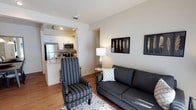 This single-floor residence offers a thoughtfully designed open-concept layout that maximizes both functionality and comfort. The living area serves as the heart of the space, featuring warm hardwood-style flooring, neutral white walls, and tasteful decorative touches including framed wall art. The room comfortably accommodates a sofa and accent chair, creating a welcoming and versatile seating arrangement centered around a patterned area rug.

Flowing directly from the living area, the kitchen is accessible via a wide archway, creating a connected yet defined space. The kitchen is equipped with stainless steel appliances, including a full-size refrigerator and microwave, complemented by white cabinetry and a stone-topped breakfast bar that doubles as a casual dining counter. Recessed lighting illuminates the kitchen workspace, while the bar seating area invites casual dining and entertaining.

The overall ambiance of the space is clean, modern, and inviting, with a cohesive neutral color palette that lends itself to easy personalization. The open sightlines between the living and kitchen areas create a sense of spaciousness, making the layout feel airy and practical for everyday living. A hallway visible from the living area suggests additional private rooms beyond, completing this well-rounded single-floor residence.