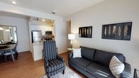 This single-floor residence offers a thoughtfully designed open-concept layout that maximizes both functionality and comfort. The living area serves as the heart of the space, featuring warm hardwood-style flooring, neutral white walls, and tasteful decorative touches including framed wall art. The room comfortably accommodates a sofa and accent chair, creating a welcoming and versatile seating arrangement centered around a patterned area rug.

Flowing directly from the living area, the kitchen is accessible via a wide archway, creating a connected yet defined space. The kitchen is equipped with stainless steel appliances, including a full-size refrigerator and microwave, complemented by white cabinetry and a stone-topped breakfast bar that doubles as a casual dining counter. Recessed lighting illuminates the kitchen workspace, while the bar seating area invites casual dining and entertaining.

The overall ambiance of the space is clean, modern, and inviting, with a cohesive neutral color palette that lends itself to easy personalization. The open sightlines between the living and kitchen areas create a sense of spaciousness, making the layout feel airy and practical for everyday living. A hallway visible from the living area suggests additional private rooms beyond, completing this well-rounded single-floor residence.
