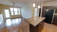 This single-floor space showcases an open-concept layout that seamlessly connects the kitchen and living areas. The kitchen is anchored by a substantial island topped with rich granite countertops, complemented by dark espresso cabinetry with sleek metal hardware. Pendant lighting hangs above the island, providing both functional task lighting and an elegant visual accent. The warm hardwood flooring extends throughout the open living area, adding a sense of continuity and warmth to the space. The living area is generously sized, bathed in natural light from multiple large windows fitted with shutters, creating a bright and welcoming atmosphere. A ceiling fan is centrally mounted overhead, enhancing comfort and airflow. Neutral-toned walls provide a clean, versatile backdrop throughout. A hallway visible in the background leads to additional rooms, suggesting a well-organized floor plan. The overall design balances functionality with a modern, refined aesthetic, making this a practical yet attractive living space suited for everyday comfort.