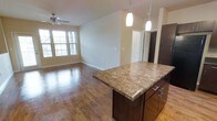 This single-floor space showcases an open-concept layout that seamlessly connects the kitchen and living areas. The kitchen is anchored by a substantial island topped with rich granite countertops, complemented by dark espresso cabinetry with sleek metal hardware. Pendant lighting hangs above the island, providing both functional task lighting and an elegant visual accent. The warm hardwood flooring extends throughout the open living area, adding a sense of continuity and warmth to the space. The living area is generously sized, bathed in natural light from multiple large windows fitted with shutters, creating a bright and welcoming atmosphere. A ceiling fan is centrally mounted overhead, enhancing comfort and airflow. Neutral-toned walls provide a clean, versatile backdrop throughout. A hallway visible in the background leads to additional rooms, suggesting a well-organized floor plan. The overall design balances functionality with a modern, refined aesthetic, making this a practical yet attractive living space suited for everyday comfort.