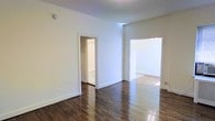 This single-floor residence presents a bright, well-maintained interior that combines classic architectural details with a clean, neutral aesthetic. The space is anchored by rich, dark hardwood floors that run throughout, adding warmth and character to each room. Crisp white walls and white trim create a fresh, airy atmosphere that enhances the sense of openness. The layout flows naturally between spaces, with wide doorways connecting the main living area to adjoining rooms. One passage leads to what appears to be a bathroom or utility area, while a wider opening connects to an additional room that benefits from generous natural light streaming in from a nearby window. A delicate chandelier is visible in the adjoining room, lending a touch of elegance to the interior. The high, smooth ceilings contribute to an expansive feel throughout. The overall ambiance is light and inviting, with a timeless, understated style that provides a versatile backdrop suited to a variety of interior arrangements and décor preferences.