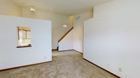 This two-story space offers a bright, open main living area characterized by soaring vaulted ceilings that lend an airy, expansive feel. The floors are covered in neutral-toned wall-to-wall carpeting, which adds warmth and comfort underfoot. A ceiling fan with a light fixture is centrally suspended, providing both illumination and ventilation to the space. The walls are finished in a clean, neutral white tone, complementing the light and versatile atmosphere throughout.

A wood-railed staircase connects the main level to the upper floor, serving as a functional architectural focal point within the room. Partial walls and architectural dividers help define distinct zones within the open layout, adding subtle structure without sacrificing the sense of openness. HVAC vents are visible in the ceiling, indicating a built-in climate control system. Dark wood baseboards provide a grounding contrast to the light walls and carpet, adding a refined finishing detail. Overall, the space presents a clean, move-in-ready environment with a functional, straightforward layout suited to a variety of uses.