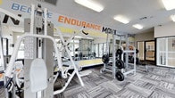 This fitness center is designed to inspire and support a comprehensive workout experience. The space is anchored by a bold, motivational mural spanning a full wall, featuring energizing phrases such as "Endurance," "Move," "Work Hard," "Progress," and "Stronger" in striking orange and yellow lettering — setting an upbeat and driven atmosphere from the moment you enter. The facility is outfitted with a robust selection of strength training equipment, including multiple cable machines, weight racks, and bench press stations loaded with plate weights, catering to a wide range of fitness levels and training styles. Large mirrors line the walls, amplifying the sense of space and allowing users to monitor their form during workouts. Overhead, recessed fluorescent lighting ensures the room is well-lit throughout. The flooring features a durable, patterned carpet tile suited for a high-traffic fitness environment. Natural light filters in through windows, adding to the bright and welcoming ambiance. The overall design of the space strikes a balance between functionality and motivation, making it a practical and energizing destination for strength and endurance training.