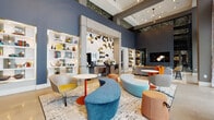 This thoughtfully designed single-floor space makes an immediate impression with its soaring ceilings and open, airy layout. The main lounge area serves as the visual centerpiece, anchored by a dramatic feature wall adorned with gold, black, and white hexagonal tiles set within a light wood frame. Built-in display shelving lines the walls on either side, backlit and styled with decorative objects and books, adding warmth and visual interest.

The lounge is furnished with an eclectic, curated collection of modern seating — including a curved blue sectional bench, swivel chairs in muted tones with bold yellow accents, and orange houndstooth ottomans — all arranged atop a large abstract area rug. The deep blue-grey wall color throughout creates a sophisticated backdrop that complements the vibrant furnishings.

A semi-private dining and meeting alcove is accessible from the main lounge, offering banquette seating with patterned cushions, white dining tables, yellow upholstered chairs, and pendant globe lighting. Deep blue accent walls and curated artwork give this nook an intimate yet stylish ambiance.

A dedicated workspace area features a wooden desk paired with a bold orange chair beside floor-to-ceiling windows dressed in abstract patterned sheer curtains, allowing ample natural light to filter through. The overall atmosphere is modern, welcoming, and design-forward.
