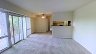 This single-floor residence offers a thoughtfully arranged open-concept layout that maximizes both comfort and functionality. The living area is a welcoming, generously sized space finished with soft, plush wall-to-wall carpeting that adds warmth underfoot. A ceiling fan with an integrated light fixture is centrally positioned overhead, providing both airflow and ambient illumination. Large sliding windows along one wall flood the interior with natural light and offer a view of the outdoors, while vertical blinds provide privacy and light control as needed.

The living space flows seamlessly into the kitchen, which is partially separated by a raised breakfast bar counter finished with a dark countertop — an ideal surface for casual dining or meal prep. The kitchen is equipped with wooden cabinetry in a warm honey tone, providing ample storage. A mirrored backsplash adds a reflective, open quality to the cooking area. The neutral, light-toned walls throughout the space complement the carpeting and create a clean, cohesive aesthetic. The overall ambiance is calm and practical, offering a functional residential environment with an approachable, move-in-ready character suitable for everyday living.