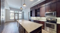 This single-floor residence combines modern design with high-quality finishes to create a refined and functional living environment. The heart of the home is an expansive gourmet kitchen, defined by rich dark-stained cabinetry extending floor to ceiling, offering abundant storage alongside open display shelving. Light granite countertops run throughout, including a generous central island illuminated by stylish pendant lighting. A full suite of stainless steel appliances — including a range, built-in microwave, dishwasher, and refrigerator — completes this chef-inspired space. Light hardwood-style flooring flows seamlessly into the open living and dining area, where soaring ceilings and oversized windows with transom glass panels flood the space with natural light and provide views of the surrounding greenery. The bedroom features plush carpeting, tall ceilings, and well-proportioned windows that maintain a bright, airy atmosphere. The bathroom is appointed with dark wood double vanity cabinetry, dual sinks with granite countertops, large framed mirrors, sconce lighting, and light-toned tile flooring, leading to a separate shower area. Throughout the residence, a cohesive palette of warm neutrals, dark wood accents, and contemporary fixtures creates a polished and inviting ambiance.