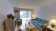 This single-floor space features a well-lit living area as its centerpiece, characterized by warm hardwood-style flooring and clean white walls that contribute to an open, airy atmosphere. A large sliding glass door dominates one wall, fitted with vertical blinds that allow for adjustable natural light while offering a direct view of the outdoors. The generous proportions of the living area comfortably accommodate a full sofa and matching armchair, both upholstered in a classic blue and white checkered pattern, arranged around a light wood coffee table. A wall-mounted air conditioning unit provides climate control, ensuring year-round comfort. A compact side table with a yellow lamp adds a warm accent to the space. Near the entrance, a white appliance, likely a fireplace or storage unit, contributes additional functionality. The overall ambiance is clean and practical, with a neutral color palette that creates a calm, welcoming environment. The abundance of natural light streaming through the sliding doors enhances the sense of space throughout the unit.
