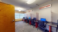 Located at 117 Washington Ave in Schenectady, NY, this single-floor professional workspace is thoughtfully designed to support both focused individual work and collaborative group sessions. The main workstation area features a row of individual computer stations equipped with desktop monitors, separated by elegant textured privacy dividers suspended from the ceiling. Bold red desk supports add a striking visual accent against the neutral tones of the walls and carpet. Decorative framed artwork panels are mounted throughout, enhancing the professional aesthetic. Natural light filters in through large windows with frosted lower panels, offering brightness while maintaining privacy. A separate meeting room provides a more intimate setting, furnished with a rounded conference table, colorful accent chairs, a wall-mounted whiteboard, and framed artwork. A soft pendant light overhead creates a warm, focused atmosphere ideal for discussions or brainstorming. Transitional open areas connect the spaces, featuring glass-paneled interior windows that allow light to flow between zones, along with potted greenery that adds a natural touch. Durable carpet tile flooring runs throughout, and warm wood-toned doors complete the cohesive, functional design of the space.
