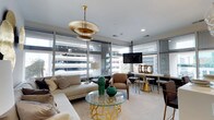 Situated in the heart of downtown Norfolk, Virginia, this thoughtfully designed single-floor space makes a strong first impression with its refined, contemporary aesthetic. Floor-to-ceiling glass windows wrap the living area, flooding the interior with abundant natural light and framing dynamic views of the surrounding urban landscape. The open layout seamlessly connects the living and dining zones, creating a cohesive and functional flow throughout the space. A striking tiered gold pendant light anchors the living area, serving as an elegant focal point above a glass-top coffee table with an ornate gold sculptural base. Neutral-toned furnishings, including a generously sized sectional sofa and taupe accent chairs, provide a warm and inviting atmosphere while complementing the overall color palette. Emerald green ceramic accents add a bold pop of color against the soft, neutral backdrop. The dining area features a sleek white table paired with stylish chairs and gold-accented bases, reinforcing the sophisticated design language found throughout. Recessed lighting and a wall-mounted flat-screen television add modern convenience, while crown molding and clean architectural lines lend a polished, finished quality to the space. Overall, the interior exudes a chic, urban sensibility with carefully curated details throughout.