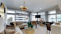Situated in the heart of downtown Norfolk, Virginia, this thoughtfully designed single-floor space makes a strong first impression with its refined, contemporary aesthetic. Floor-to-ceiling glass windows wrap the living area, flooding the interior with abundant natural light and framing dynamic views of the surrounding urban landscape. The open layout seamlessly connects the living and dining zones, creating a cohesive and functional flow throughout the space. A striking tiered gold pendant light anchors the living area, serving as an elegant focal point above a glass-top coffee table with an ornate gold sculptural base. Neutral-toned furnishings, including a generously sized sectional sofa and taupe accent chairs, provide a warm and inviting atmosphere while complementing the overall color palette. Emerald green ceramic accents add a bold pop of color against the soft, neutral backdrop. The dining area features a sleek white table paired with stylish chairs and gold-accented bases, reinforcing the sophisticated design language found throughout. Recessed lighting and a wall-mounted flat-screen television add modern convenience, while crown molding and clean architectural lines lend a polished, finished quality to the space. Overall, the interior exudes a chic, urban sensibility with carefully curated details throughout.