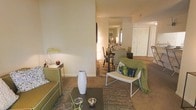 This single-floor residence offers a thoughtfully designed open-concept layout that seamlessly connects the living and dining areas, creating a sense of spaciousness and flow throughout. The living area features warm, neutral-toned carpeting and is furnished with a comfortable sofa, a sleek dark coffee table, and a modern accent chair draped in a cozy green throw — all anchored by a textured area rug that adds depth and warmth. Decorative touches such as a white ceramic vase, metallic accent bowls, and a green desk lamp contribute to the space's curated, contemporary aesthetic. A partial wall subtly divides the living space from the dining area, which is visible in the background and features a full dining table with seating for multiple guests, illuminated by warm ambient lighting. The neutral palette of soft whites, beiges, and earthy greens is carried consistently throughout, lending the interior a calm and cohesive feel. Built-in storage and closet space are also visible, enhancing the functional appeal of the layout. Natural light filters into the space, complementing the warm artificial lighting to create an inviting and comfortable atmosphere suited for both relaxed living and entertaining.