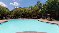 This amenity complex offers two distinct recreational spaces designed to support an active lifestyle. The outdoor swimming pool is a generously sized, freeform aquatic retreat with clear turquoise water, surrounded by a spacious pool deck finished in warm-toned pavers. Lounge chairs and umbrella-shaded seating are arranged around the perimeter, inviting relaxation in the open air. Mature trees frame the background, providing natural shade and a lush, park-like setting beneath open skies.

The fitness center is thoughtfully divided into multiple interconnected zones, each tailored to different workout styles. The main exercise area features rubber flooring, a large wall-mounted smart TV for on-demand fitness programming, and an assortment of free weights and stability balls. Interior windows connect the spaces visually, creating an open, collaborative feel while allowing natural light to filter through. A dedicated cycling studio houses multiple stationary spin bikes alongside a mirrored wall and a window overlooking the outdoors. Additional areas include a cardio zone with machines such as ellipticals and treadmills, resistance bands mounted to the wall, and a ductless mini-split climate control system ensuring year-round comfort throughout the facility.