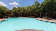 This amenity complex offers two distinct recreational spaces designed to support an active lifestyle. The outdoor swimming pool is a generously sized, freeform aquatic retreat with clear turquoise water, surrounded by a spacious pool deck finished in warm-toned pavers. Lounge chairs and umbrella-shaded seating are arranged around the perimeter, inviting relaxation in the open air. Mature trees frame the background, providing natural shade and a lush, park-like setting beneath open skies.

The fitness center is thoughtfully divided into multiple interconnected zones, each tailored to different workout styles. The main exercise area features rubber flooring, a large wall-mounted smart TV for on-demand fitness programming, and an assortment of free weights and stability balls. Interior windows connect the spaces visually, creating an open, collaborative feel while allowing natural light to filter through. A dedicated cycling studio houses multiple stationary spin bikes alongside a mirrored wall and a window overlooking the outdoors. Additional areas include a cardio zone with machines such as ellipticals and treadmills, resistance bands mounted to the wall, and a ductless mini-split climate control system ensuring year-round comfort throughout the facility.