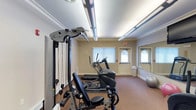 This single-level fitness room is thoughtfully designed to accommodate a variety of exercise routines. The space is outfitted with a Hoist multi-station strength training machine, providing options for a comprehensive resistance workout. Complementing the strength equipment, a Vision Fitness elliptical and a stair climber offer effective cardio training options. A large wall-length mirror spans one side of the room, enhancing the sense of space while allowing users to monitor their form during workouts. A stability ball adds a functional training element to the room.

The room features carpeted flooring in a warm earth tone, providing comfort underfoot. Crown molding trims the ceiling, lending a refined touch to the space. Overhead, long fluorescent light panels provide consistent, bright illumination throughout the room. Natural light filters in through two windows dressed with horizontal blinds, while a set of doors with arched window details at the far end of the room offers both an exit point and additional natural light. The overall ambiance is clean, functional, and well-maintained.