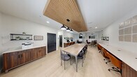 This business center is a thoughtfully designed, single-floor workspace that efficiently combines multiple functional areas within a compact footprint. The interior is defined by a clean, modern aesthetic, featuring light wood-look flooring, crisp white walls, and a striking slatted wood ceiling detail that adds warmth and texture overhead.

The central conference and dining area accommodates a large rectangular table surrounded by upholstered chairs, illuminated by a trio of industrial-style pendant lights. Adjacent to this area, a dedicated workstation zone lines the wall with a continuous desk surface, walnut-toned cabinetry, dual computer monitors, and open shelving decorated with small plants—providing an organized workspace for individual tasks.

A printing and storage station is conveniently located nearby, featuring a built-in cabinet with a printer and wall-mounted shelving. Dark-paneled doors lead to a private meeting room enclosed by glass partitions, furnished with a round table, leather chairs, and a wall-mounted display screen—ideal for small group discussions or video conferencing. The overall ambiance is professional yet inviting, balancing productivity-focused functionality with polished, contemporary design details throughout.