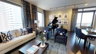 This single-floor space presents a thoughtfully curated living area that blends elegance with comfort. The room centers around a striking black baby grand piano, which serves as a dramatic focal point and immediately signals a refined sensibility. Surrounding it, the seating arrangement includes a cream-toned sofa and a pair of deep navy tufted armchairs, all accented with navy and white geometric throw pillows that echo the bold patterned drapery framing the large windows. The drapes, featuring an eye-catching lattice print in navy and white, extend from floor to ceiling and contribute to a sense of height and grandeur. Natural light filters in generously through the windows, brightening the space and complementing the warm-toned hardwood floors beneath. A richly hued blue-toned area rug anchors the seating zone, adding depth and texture to the overall palette. A dark wood coffee table with a decorative tray, fresh floral arrangement, and books adds a lived-in, welcoming touch. Along one wall, a curated gallery of framed black-and-white photographs lends an artistic, gallery-like quality. Track lighting overhead provides controlled illumination, enhancing the space's polished and inviting ambiance.