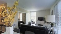 This single-floor residence showcases a thoughtfully designed interior that blends modern aesthetics with comfortable living. The open-concept layout creates a seamless flow between the living and dining areas, making the space feel expansive and inviting.

The living area is anchored by a sleek black leather sofa and a striped area rug, complemented by a wicker accent chair positioned near a large window dressed with horizontal blinds. Natural light filters generously into the room, brightening the neutral, warm-toned walls and smooth white ceilings. A minimalist white media console provides storage and display space, paired with a flat-screen television.

Decorative touches, including a vibrant yellow flowering branch arrangement and potted greenery, introduce natural elements that enliven the monochromatic color scheme. A round black side table adds visual contrast, while framed wall art contributes a curated, gallery-like quality to the space.

An adjacent room, visible through an open doorway, suggests additional living or sleeping quarters, reinforcing the residence's functional layout. The dining area, partially visible in the foreground, offers seating with modern dark chairs. Overall, the space exudes a clean, contemporary ambiance that is both practical and visually sophisticated.