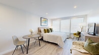 Located at 50 Morgan Street in Hartford, Connecticut, this thoughtfully designed studio unit offers a compact yet functional single-floor layout with one bathroom and a dedicated entrance. The main living area is bright and open, featuring light-toned hardwood-style flooring, recessed lighting, and large windows with blinds that flood the space with natural light. The room comfortably accommodates a sleeping area, a cozy seating nook with a loveseat accented by a decorative sunburst mirror, and a small dining table — all flowing seamlessly together. A kitchenette lines one wall, equipped with white cabinetry, a stainless steel refrigerator, a microwave, and a dark-toned countertop with a sink, providing practical cooking and storage solutions. Wall art with bold, modern colors adds a lively visual accent to the neutral white walls. The bathroom is cleanly finished with large-format gray tile flooring, a warm wood vanity with a white basin, matte black fixtures, and a lit mirror, creating a polished and functional space. The overall ambiance is fresh, modern, and welcoming — well-suited for efficient single-occupancy living in the Hartford area.
