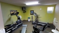 This single-floor fitness room provides a practical and functional space for exercise. The room features yellow-green painted walls and a textured ceiling with overhead fluorescent lighting that brightens the interior. The carpeted floor adds a layer of comfort underfoot throughout the space.

The room is equipped with a selection of cardio equipment, including an elliptical trainer, a stationary recumbent bike, and a treadmill, allowing users to engage in a variety of aerobic workouts. A flat weight bench is also available for strength training exercises. A tower fan is positioned in the room to provide air circulation during workouts.

For entertainment, a wall-mounted CRT television with an accompanying media device is installed in the corner, allowing users to watch TV while exercising. A framed wall print adds a modest decorative touch to the space. A round analog clock is mounted on one of the walls, keeping users informed of the time during their sessions.

Dumbbell weights are neatly stored on a small stand, offering additional options for resistance training. Overall, the fitness room is a well-organized, no-frills space designed to meet the essential workout needs of its users.