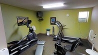 This single-floor fitness room provides a practical and functional space for exercise. The room features yellow-green painted walls and a textured ceiling with overhead fluorescent lighting that brightens the interior. The carpeted floor adds a layer of comfort underfoot throughout the space.

The room is equipped with a selection of cardio equipment, including an elliptical trainer, a stationary recumbent bike, and a treadmill, allowing users to engage in a variety of aerobic workouts. A flat weight bench is also available for strength training exercises. A tower fan is positioned in the room to provide air circulation during workouts.

For entertainment, a wall-mounted CRT television with an accompanying media device is installed in the corner, allowing users to watch TV while exercising. A framed wall print adds a modest decorative touch to the space. A round analog clock is mounted on one of the walls, keeping users informed of the time during their sessions.

Dumbbell weights are neatly stored on a small stand, offering additional options for resistance training. Overall, the fitness room is a well-organized, no-frills space designed to meet the essential workout needs of its users.