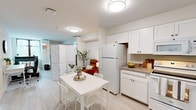 Located at 1698 University Ave in Berkeley, CA, this efficiently designed single-floor unit offers a clean, modern aesthetic defined by crisp white walls, light wood-style flooring, and flush ceiling lighting throughout. The open-plan living and kitchen area forms the heart of the space, where white shaker-style cabinetry, a full-size refrigerator, and a compact countertop workspace provide a fully functional kitchen. A dining area with a white table and chairs sits adjacent, creating a seamless flow between cooking and dining. A decorative lattice-panel room divider elegantly defines the transition into the living zone, adding visual interest without closing off the open feel. The spacious primary bedroom features a large window with a dark-framed surround that invites natural light and offers outdoor views, complemented by warm neutral walls and ample room for both rest and a work setup. A dedicated office nook, separated by the same stylish room divider, includes built-in shelving and space for a full desk configuration — ideal for remote work or study. The bathroom is clean and functional, featuring a wall-mounted sink, mirrored medicine cabinet, and light, airy finishes. The overall ambiance is minimal, bright, and practical, well-suited for urban living in the heart of Berkeley.