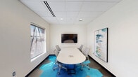 This single-floor office suite offers a well-organized and thoughtfully designed workspace that balances functionality with a contemporary aesthetic. The main area is divided into two distinct zones: a collaborative conference area and an open workstation section. The conference area is anchored by a sleek, oval white table surrounded by ergonomic mesh chairs, a wall-mounted flat-screen display, and framed artwork, creating a polished environment for meetings and presentations. Two windows provide generous natural light and elevated city views, enhancing the professional atmosphere. The workstation section features a long, continuous white desk running along the wall, accompanied by additional ergonomic seating and wall-mounted trailing plants that add a refreshing, biophilic element to the space. An HP printer station is also integrated into this area for added convenience. Throughout the main space, a striking hexagonal carpet tile pattern in shades of blue, teal, and gray adds visual energy and a modern design sensibility. The suite also includes a private bathroom finished with gray stone-look floor and wall tiles, a wall-mounted sink, a framed mirror, and ADA-compliant grab bars and fixtures. Recessed lighting and a dropped ceiling grid are consistent throughout, contributing to a clean, professional finish.