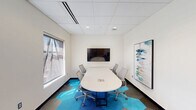 This single-floor office suite offers a well-organized and thoughtfully designed workspace that balances functionality with a contemporary aesthetic. The main area is divided into two distinct zones: a collaborative conference area and an open workstation section. The conference area is anchored by a sleek, oval white table surrounded by ergonomic mesh chairs, a wall-mounted flat-screen display, and framed artwork, creating a polished environment for meetings and presentations. Two windows provide generous natural light and elevated city views, enhancing the professional atmosphere. The workstation section features a long, continuous white desk running along the wall, accompanied by additional ergonomic seating and wall-mounted trailing plants that add a refreshing, biophilic element to the space. An HP printer station is also integrated into this area for added convenience. Throughout the main space, a striking hexagonal carpet tile pattern in shades of blue, teal, and gray adds visual energy and a modern design sensibility. The suite also includes a private bathroom finished with gray stone-look floor and wall tiles, a wall-mounted sink, a framed mirror, and ADA-compliant grab bars and fixtures. Recessed lighting and a dropped ceiling grid are consistent throughout, contributing to a clean, professional finish.