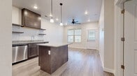 This single-floor residence offers a thoughtfully designed open-concept layout where the kitchen flows seamlessly into the living area, creating a cohesive and functional living space. The kitchen is a standout feature, showcasing rich dark-brown cabinetry paired with light granite countertops that extend across both the perimeter workspace and a central island. Stainless steel appliances, including a refrigerator, range, microwave, and dishwasher, complement the modern aesthetic, while open floating shelves add a contemporary touch alongside the upper cabinetry. Pendant lighting above the island and recessed ceiling lights provide layered illumination throughout the space. Light wood-look flooring runs consistently throughout, lending warmth and visual continuity from room to room. The living area benefits from natural light through blinded windows and is equipped with a ceiling fan for year-round comfort. A private exterior door connects directly to an outdoor area. The bathroom maintains the same design language, featuring a granite-topped dark vanity with an undermount sink, a large mirror, wood-look tile flooring, and a full tub-shower combination. Overall, the space presents a clean, modern aesthetic with cohesive finishes throughout.