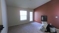This single-floor space centers around a comfortable living area distinguished by a warm terracotta-colored accent wall that creates a focal point and adds character to the otherwise neutral interior. The remaining walls are finished in a clean, light beige tone, contributing to a bright and airy atmosphere. The room is carpeted in a soft, neutral-toned material that extends throughout, providing a comfortable underfoot feel. Natural light filters generously into the space through a large window and a glass-paneled exterior door positioned side by side along one wall, offering potential outdoor access and allowing daylight to illuminate the interior. The exterior door features decorative glass panels, adding a subtle architectural detail. Standard white baseboards frame the perimeter of the room, maintaining a clean and cohesive look. A small decorative display, including a chalkboard-style frame and accent pillows, is positioned near the accent wall, hinting at the space's versatile potential for personalization. The overall ambiance is warm and welcoming, with a straightforward layout that lends itself well to flexible furniture arrangements and everyday living.