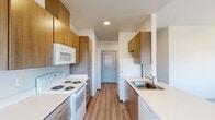 Located in Salem, Oregon, this efficiently designed one-bedroom, one-bathroom unit offers a well-organized single-floor layout. The galley-style kitchen and dining area are finished with warm wood-toned cabinetry extending to the ceiling, white quartz-style countertops, and wood-look plank flooring. White appliances including a coil-top electric range, over-range microwave, refrigerator, and dishwasher equip the kitchen fully. Natural light filters in through a window at the dining end, creating a bright and open feel in the cooking and eating space. The living room features carpet flooring, multiple windows that bring in generous natural light, and a glass-paneled exterior door that provides views of the surrounding neighborhood. A wall-mounted HVAC unit serves the living area. The bedroom is carpeted and comfortably proportioned, offering a reach-in closet with a hanging rod and a window with outdoor views. An additional closet provides supplementary storage. The combined bathroom and laundry room is a practical highlight, housing a full-size washer and dryer, a toilet, towel bars, overhead shelving, and a vanity with cabinetry — all finished with wood-look flooring and a small window for ventilation. The clean, neutral palette throughout gives the unit a fresh, move-in-ready appearance.
