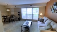 This single-floor space offers a warm and inviting atmosphere designed for comfortable everyday living. The living area serves as the centerpiece of the home, featuring a large bank of triple windows that flood the room with natural light and offer pleasant views of the surrounding greenery outdoors. Plush wall-to-wall carpeting adds a soft, cozy underfoot feel throughout, while neutral-toned walls provide a versatile backdrop that complements a variety of décor styles. The room is well-proportioned, providing ample space for a full seating arrangement, including a sofa and a glass-top coffee table, making it ideal for both relaxing and entertaining. Decorative touches such as potted topiary plants add a tasteful, natural element to the space. A dining area is visible nearby, seamlessly connected to the living space to create an open, flowing layout. Ceiling vents throughout ensure consistent climate control year-round. The overall ambiance is clean, bright, and functional, with a classic residential character that offers a comfortable and practical living environment suitable for a range of lifestyles.