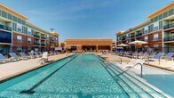 Located at 321 E Park Rd in Indian Trail, North Carolina, this outdoor amenity space is designed to deliver a resort-style experience within a residential community. At its centerpiece is a generously sized rectangular swimming pool with lane markings, flanked by rows of lounge chairs and sun umbrellas that invite relaxation under the open sky. The pool area is framed by multi-story brick and siding residential buildings, creating an enclosed, community-focused atmosphere.

Adjacent to the pool is a covered outdoor lounge area that serves as a social hub. The space features coffered ceilings with decorative windmill-style ceiling fans, open panel walls in sage green and white, and a built-in fireplace at its rear. Comfortable seating arrangements with navy blue cushions and lime green accent pillows are paired with sleek low-profile tables, offering a welcoming setting for gathering year-round.

The surrounding walkways are finished in clean concrete and lined with manicured landscaping, mulched beds, and ornamental black metal fencing. Wall-mounted lantern-style light fixtures add a classic touch along the brick building facades. Accessible amenities such as a dual water fountain station are thoughtfully integrated into the exterior walls. The overall design balances functional outdoor living with an attractive, well-maintained aesthetic suited to community use.